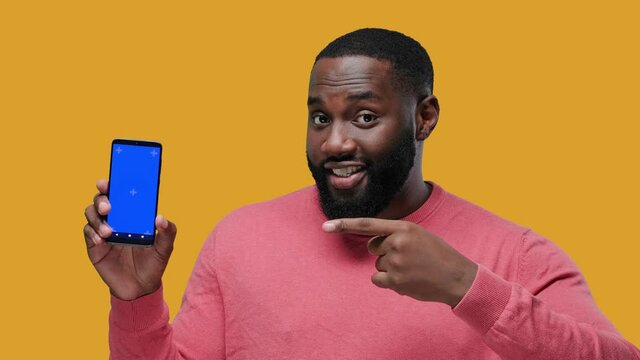 Happy young black guy showing his phone screen with chroma key and pointing to it on yellow background