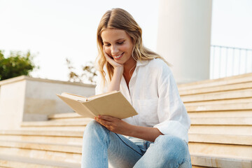 Beautiful happy woman smiling and reading book while sitting on staircase