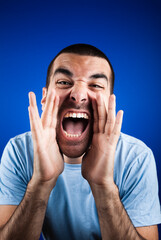 Dark short haired man in light blue t shirt, holding hands around his mouth, screaming. Studio photo, blue background.