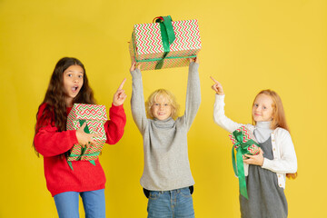 Giving and getting presents on Christmas holidays. Group of happy smiling children having fun, celebrating isolated on yellow studio background. New Year 2021 meeting, childhood, happiness, emotions.