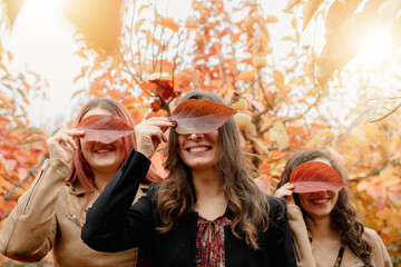 Three girls portrait in a field smiling. Close up covering eyes with a red leaf. Autumn, friendship concept.