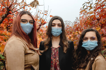 Three girls portrait looking at the camera wearing protective face mask from Coronavirus infection. Autumn background, red fallen leaves, social distancing and pandemic concept.