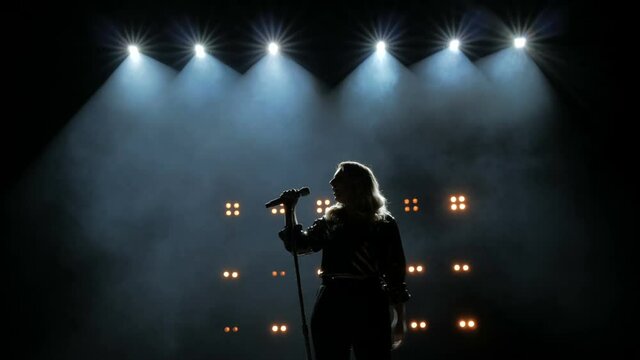 Silhouette of a girl comes to the microphone on the stage against a background of bright yellow lights