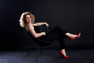 Young blond curly woman, sitting on chair in front of black background. Full-length portrait of female, wearing black clothes and red high heels. Love yourself concept. Studio dark key photography.
