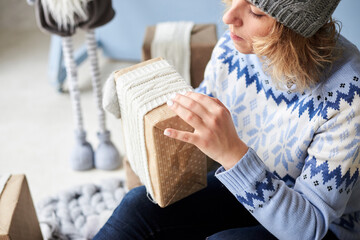 Young woman, wearing light blue sweater with norwegian swedish pattern is about to open christmas present in light room with holiday decorations. Merry Christmas and Happy new year. Winter holidays