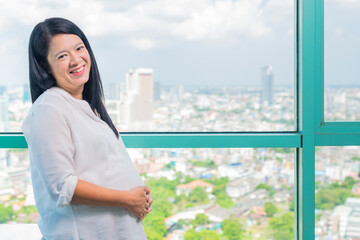 Fototapeta premium Asian pregnant woman touching her belly with hands and thinking of future. She taking a break pensive standing near condo window.