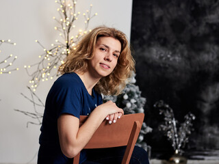 Young blond curly woman, wearing blue t-shirt, sitting in front of decorated wall with christmas...