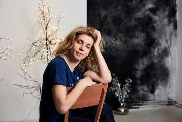 Fototapeta na wymiar Young blond curly woman, wearing blue t-shirt, sitting in front of decorated wall with christmas trees and lights, leaning on her head. Merry christmas, happy new year celebration party.