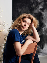 Fototapeta na wymiar Young blond curly woman, wearing blue t-shirt, sitting in front of decorated wall with christmas trees and lights, leaning on her head. Merry christmas, happy new year celebration party.