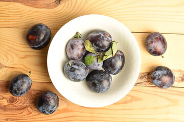 Ripe dark blue organic plums in a ceramic bowl, close-up, on a wooden table.