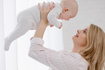 A young Caucasian mother in home clothes holds a newborn baby in a white jumpsuit. Smiles and hugs the child.