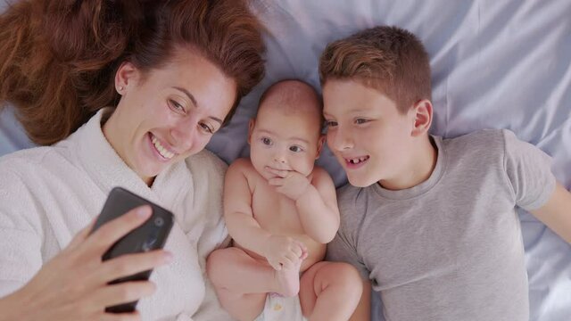 Happy mother with her kids are making a selfie or video call to father or relatives in a bed. Concept of technology, new generation,family, connection, parenthood, authenticity.