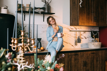 Charming brunette in light blue pyjamas with coffee and croissant in kitchen in Christmas morning.