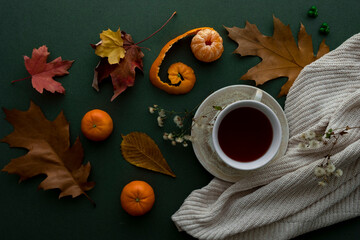 Cup of tea with tangerines, fall leaves and sweaters on green background. Cosy autumn-winter concept.