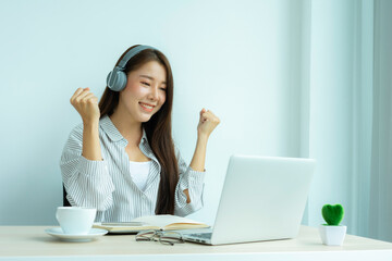 A young Asian woman who looks happily wears headphones. Watch webinars, listen to online courses, communicate by video conferences and learn languages, laughing, studying with teacher lessons via vide