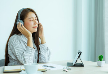 side view portrait of a beautiful Asian woman wearing headphones smiling and closing her eyes to listen to the music played by the tablet. At her desk in the home office after work is over, the work-f