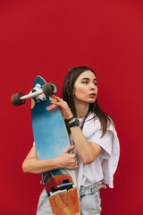 Portrait of stylish woman in casual clothes standing on red background with longboard in hands,...
