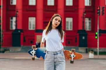 Stylish female skater in stylish casual clothes stands on the street and poses with a gongboard behind her back, looking at the camera with a serious face. Vertical © bodnarphoto