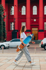 Stylish woman with a longboard in her hands walks down the city street with a serious face....
