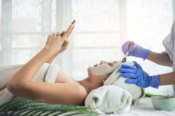 Fototapeta premium Asian woman relax use smartphone and getting facial face care by beautician at spa salon.