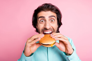 Photo portrait of hungry man taking massive bite of cheeseburger isolated on pastel pink colored background