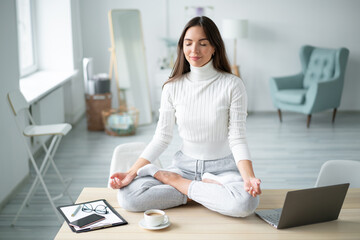 Young woman sitting with closed eyes desk next to laptop in padmasana yoga pose home office.