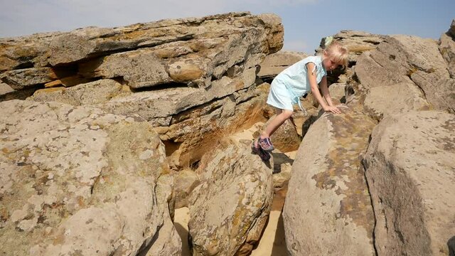 The child climbs stones and stones. Sand is visible between the stones. Nice summer day. The girl enjoys overcoming obstacles. A girl with blond hair in a light white dress and sandals. 