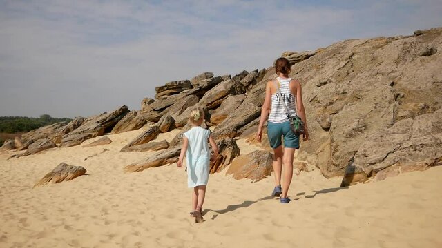 Mom and child climb the stones and rocks, sand is visible between the stones. Nice summer day. The family enjoys overcoming obstacles. A girl with blond hair in a light white dress and sandals. 