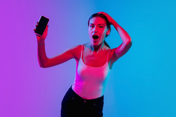 Showing phone screen. Young caucasian woman's portrait on gradient blue-purple studio background in neon light. Concept of youth, human emotions, facial expression, sales, ad. Beautiful brunette model