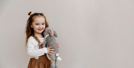 portrait of a little cute girl with a toy in her hands on a beige background. Three year old child smiling and looking at the camera. handmade knitted toy hare