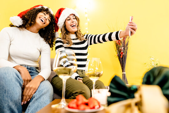 Family Holidays With Diversity: Beautiful Blonde Takes A Selfie At Home With Her Black Hispanic Girlfriend Wearing Santa Hat - Modern People Different Relationships And Habits With Technology