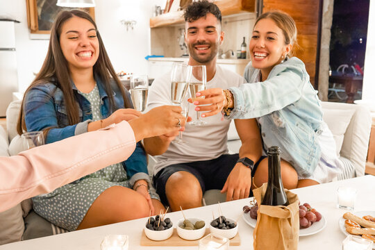 Young friends having fun toasting with champagne at home with aperitif snacks on the table - Prosecco wine to forge bonds between people that want to enjoy their life - Focus on glasses