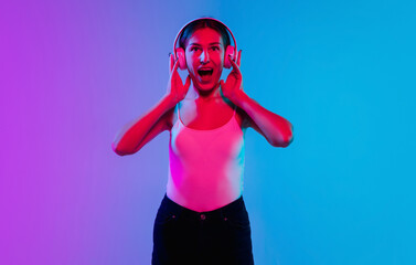 Listening to music with headphones. Young caucasian woman's portrait on gradient blue-purple studio background in neon. Concept of youth, human emotions, facial expression, sales, ad. Beautiful model.