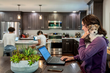 Woman working from home on laptop computer holding cute puppy while daughters study at kitchen counter in background