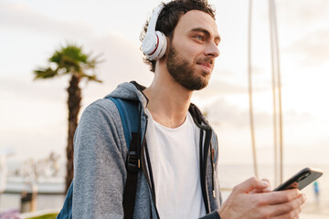 Pleased young guy using mobile phone while walking on promenade