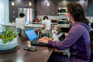 Woman working from home on laptop computer with puppy in lap while daughters study at kitchen counter in background