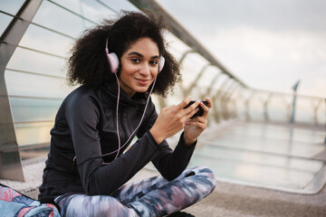 Cheerful athletic sportswoman in headphones using mobile phone