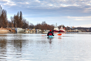 Fototapeta na wymiar Man kayaking on blue kayak in winter river at the evening. View on town