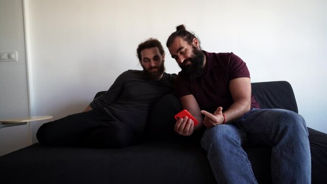 One bearded man shows to his friend a mobile app at home while both are sitting on a sofa at the living room