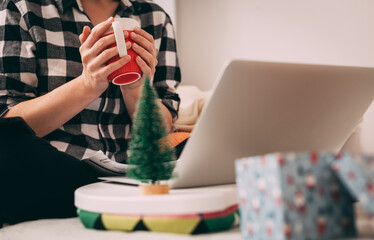 Woman in plaid shirt  sitting on bed in front of laptop and holding Christmas mug in her hands