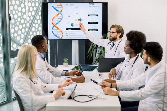 Waist up portrait of multi-ethnic five doctors on medical staff meeting, looking at the digital screen, while sitting at table and discussing. Male Caucasian doctor, showing his colleagues the chart