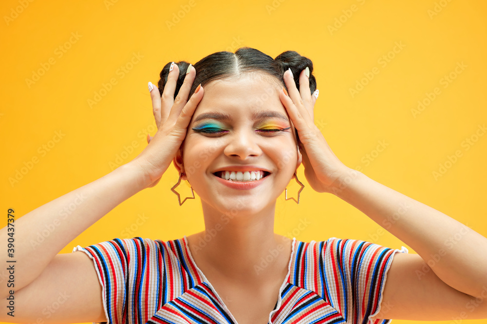 Wall mural young woman with colorful eyes smiles and covers her face with her hands, posing on yellow backgroun