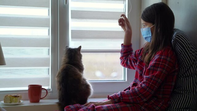 Girl In Pajamas Wearing Medical Mask Sitting Near Window And Opening Blinds, Cat Watching The Process. Sick Child Staying At Home At Quarantine And Spending Time With Pet. Concept Of Healthcare