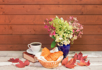 Cup of coffee, croissants, book and autumnal fading pink and white hydrangea flowers in vase on aged wooden table. View with copy space.