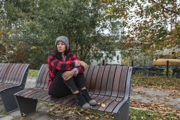 Young depressed homeless girl or woman with hat sitting alone on the bench on the street in the...