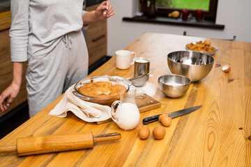Tasty apple pie on a oaken wood table with a ingredients. Woman in a gray pajama cooking a delicious apple pie on a kitchen in a loft style. Milk, eggs, sugar and a baked pie on a table. Home kitchen.