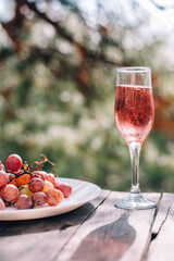 A glass of champagne or wine next to pink grapes is placed on a wooden table against a background of pine and juniper forests