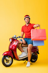 happy delivery man in helmet sitting on scooter and holding shopping bags on yellow