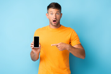 shocked man in t-shirt pointing with finger at smartphone with blank screen on blue