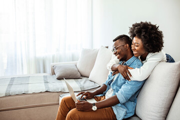 Naklejka na ściany i meble Happy young african couple with laptop computer. Happy black couple working together on laptop sitting on sofa at home. Freelance and remote work concept, copy space